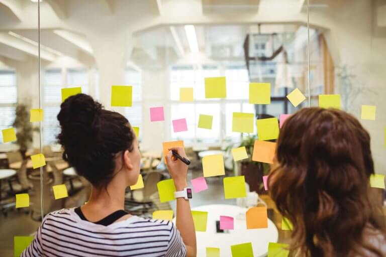 two-women-writing-on-sticky-notes-attached-to-glass